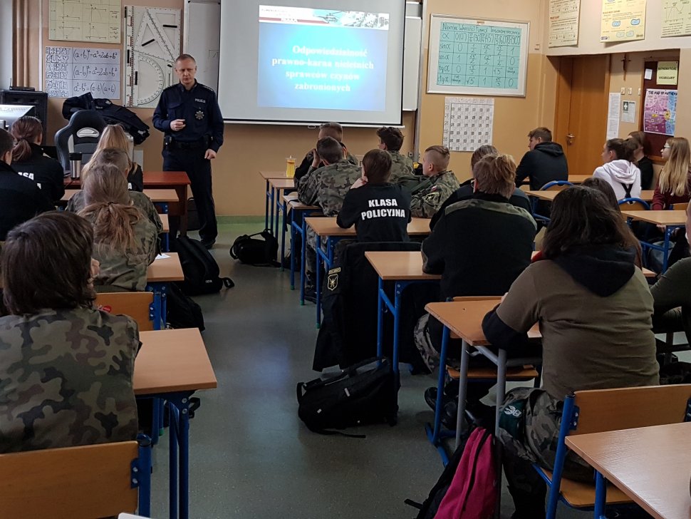 Policjant (stoi na drugim planie) na prelekcji w sali lekcyjnej PZS w Lędzinach. Uczniowie siedzą w ławkach.