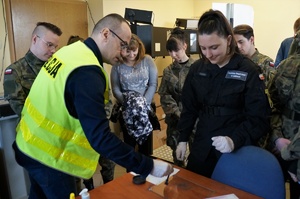 Zajęcia u technika kryminalistyki. Policjant w odblaskowej kamizelce prezentuje uczniom techniki pobierania śladów. Uczniowie w mundurach stoją wokół policjnata