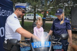 Na policyjnym motocyklu siedzi mała dziewczynka. Po lewej stronie stoi policjant ruchu drogowego, po prawej policjantka z komendy w Bieruniu. W tle widać drzewa