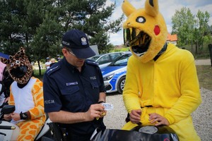 Policjant stoi przy motocykliście w żółtym stroju bajkowej postaci wręczając mu profilaktyczny gadżet. W tle widać drzewa