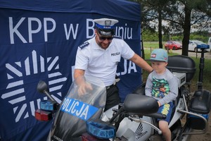 Na tle profilaktycznego namiotu, na którym widoczne jest logo Policji, przy służbowym motocyklu stoi policjant ruchu drogowego. Na motocyklu siedzi mały chłopiec
