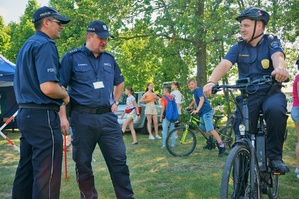 W lewej części zdjęcia stoją dwaj policjanci, w tym profilaktyk i Komendant Powiatowy Policji w Bieruniu. W prawej części Komendant Straży Miejskiej w Bieruniu siedzi na rowerze. W tle widać drzewa i uczestników festynu