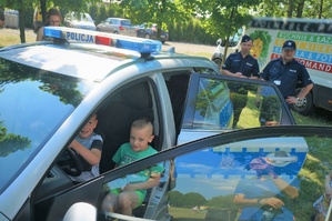 W policyjnym radiowozie za kierownicą siedzi dwóch małych chłopców. Za radiowozem stoją dwa policjanci. W tle widać drzewa