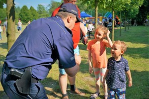 Policjant po lewej stronie nachyla się nad dwójką małych dzieci rozmawiając z nimi. W tle widać boisko Mini Arboretum