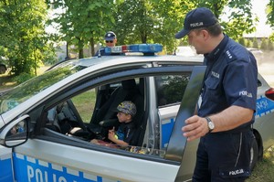 Komendant Powiatowy Policji w Bieruniu stoi przy radiowozie trzymając uchylone drzwi. Za kierownicą siedzi dziecko. W tle widać drzewa