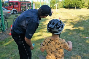 Policjant nachyla się nad chłopcem w kasku ochronnym siedzącym na rowerku wskazując palcem na rowerowy tor przeszkód, który widać na drugim planie