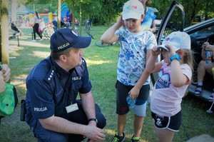 Na trawniku przy radiowozie Komendant Powiatowy Policji w Bieruniu kuca przed dwójką małych dzieci rozmawiając z nimi